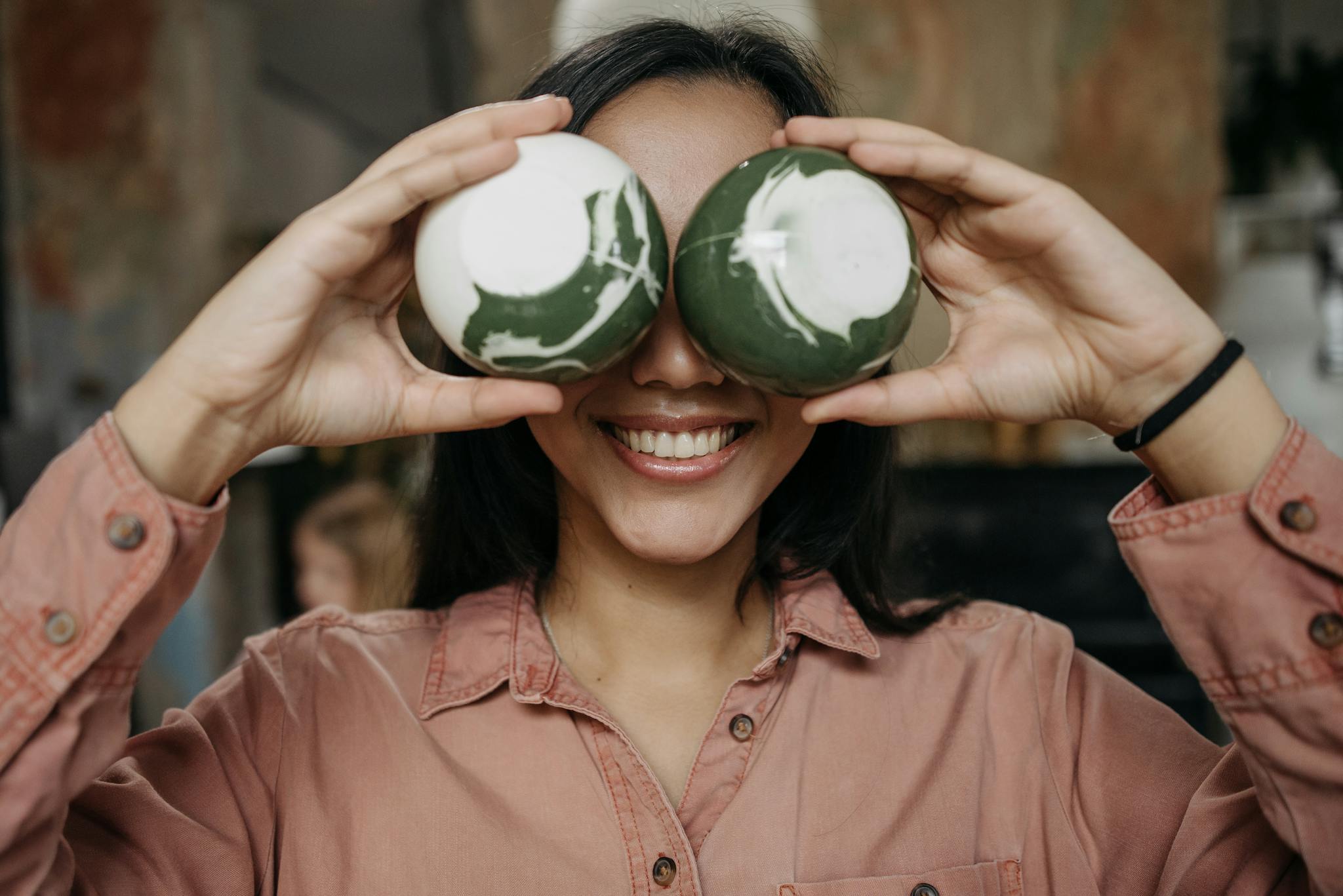 Cheerful woman using ceramic bowls to cover her eyes in a playful indoor setting.